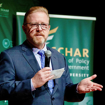 A man in a blue suit stands in front of a green background and holds a microphone.