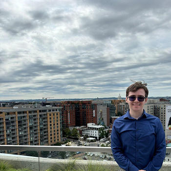A young man wearing a blue shirt and sunglasses stands in front of a glass fence overlooking Washington, D.C., with the U.S. Capitol in the distance.