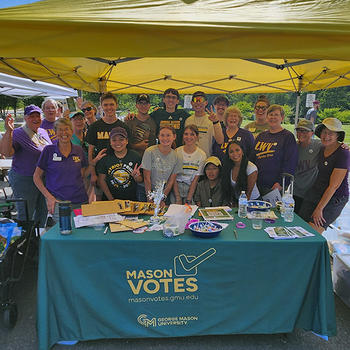 A large group of students and community volunteers stand smiling under a yellow tent behind a Mason Votes information table filled with registration materials.
